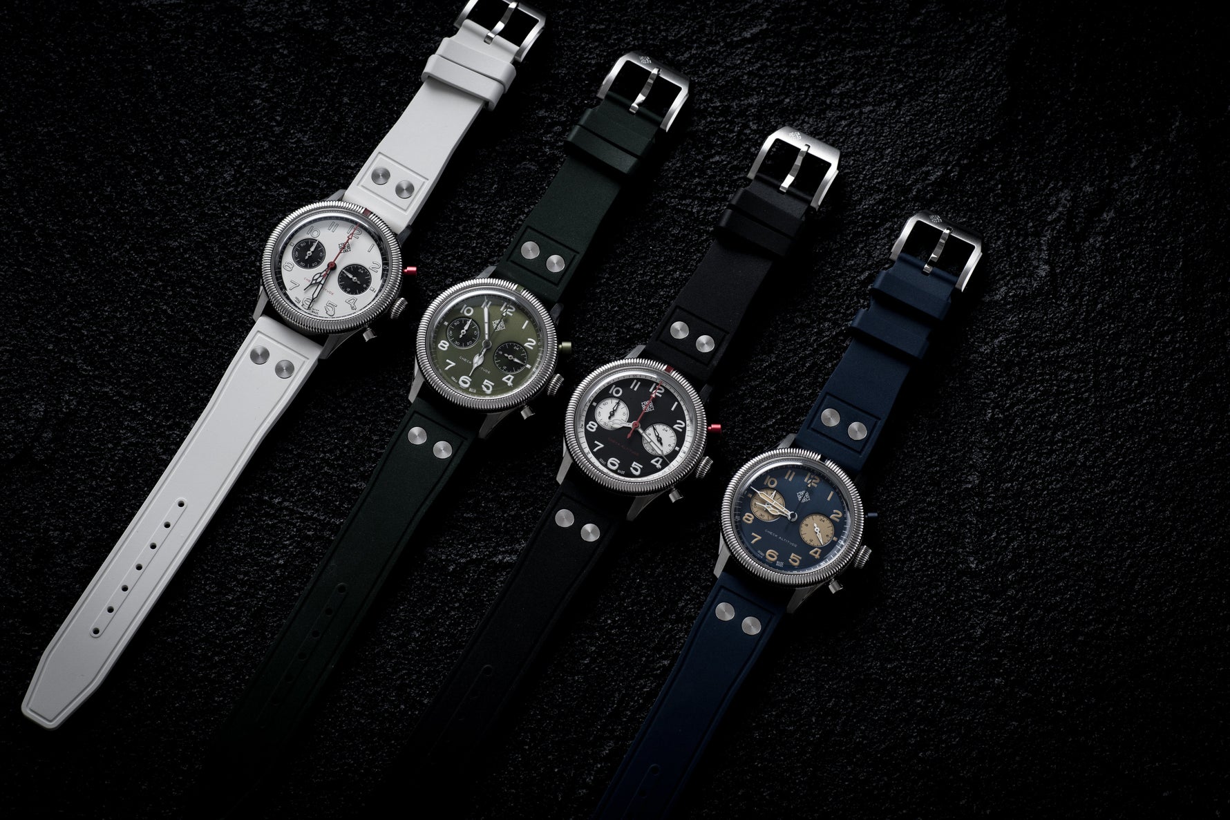 Four IXDAO Pilot Chronograph watches in white, green, black, and blue colorways, displayed diagonally on a textured black background.